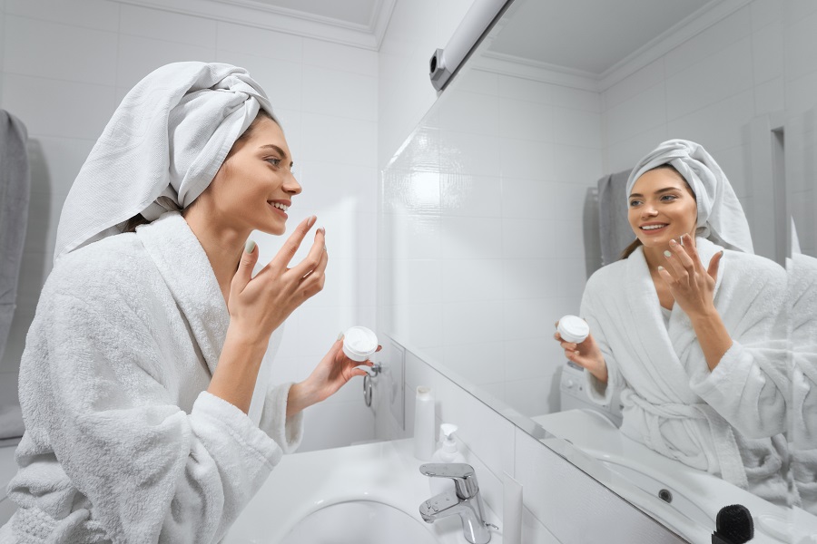 Side view of smiling cute young woman apply facial cream look in mirror. Concept of body care and preparing skin for makeup with good mood at home or hotel.